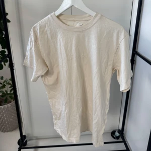 Beige t-shirt från (di)vision - Säljer en stilren beige t-shirt från (di)vision med tryck på baksidan. T-shirten har en normal passform och är tillverkad i ett mjukt material. Perfekt för en avslappnad look.