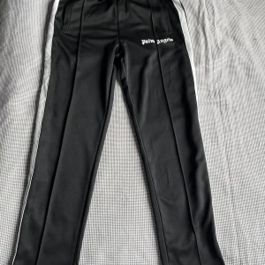 Palm Angels byxor  - Snygga svarta träningsbyxor från Palm Angels med vita sidoremsor. Byxorna har en elastisk midja och logotypen tryckt på framsidan. Perfekta för en sportig look.