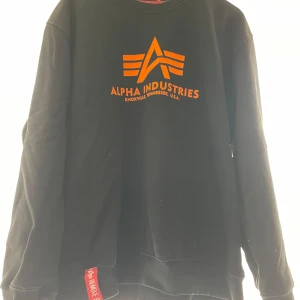 Svart sweatshirt från Alpha Industries - Säljer en svart sweatshirt från Alpha Industries med orange logga på framsidan. 