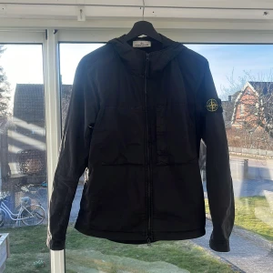 Svart jacka från Stone Island - Säljer en stilren svart jacka från Stone Island med huva och dragkedja. Jackan har en broderad logotyp på ärmen och är perfekt för en casual look. Den är långärmad och har en modern design. Köpt på NK i Gbg.
