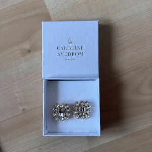 Glittriga örhängen från Caroline Svedbom - Snygga glittriga örhängen från Caroline Svedbom. De har en elegant design med klara stenar i en blomliknande form. Perfekta för att ge en touch av glamour till din outfit.
