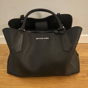 Svart handväska från Michael Kors - Snygg svart handväska från äkta Michael Kors i skinn med stilren design. Väskan har två handtag och en praktisk dragkedja upptill. Perfekt till vardags. Kostar originellt ca 2500kr. Köpt i LA. Pris kan diskuteras 