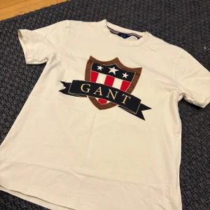 Vit t-shirt från GANT - Säljer en stilren vit t-shirt från GANT med ett stort tryck av deras ikoniska logga på framsidan. T-shirten har korta ärmar och en klassisk rund halsringning. Perfekt för en avslappnad look!