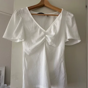 Vit ribbad blus med v-ringning från Vero Moda - Säljer en stilren vit blus med v-ringning och från Vero Moda. Blusen har korta ärmar och är tillverkad i ett ribbat material som ger en fin struktur. Perfekt för både vardag och festliga tillfällen.