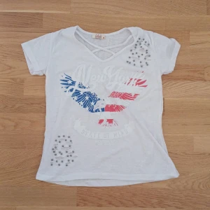 Vit t-shirt med New York-tryck från Caliente - Säljer en vit t-shirt från Caliente med ett coolt New York-tryck i rött och blått. T-shirten har korta ärmar och är dekorerad med silverfärgade pärlor. Perfekt för en avslappnad stil!