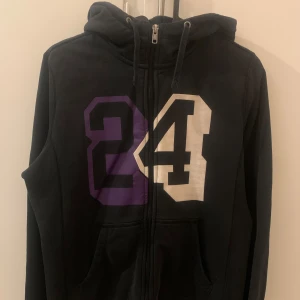 Nike hoodie - Säljer en svart hoodie från Nike med ett stort tryck av siffran 24 i lila och vitt på framsidan. Hoodien har dragkedja och en praktisk huva med dragsko. Perfekt för en avslappnad stil.