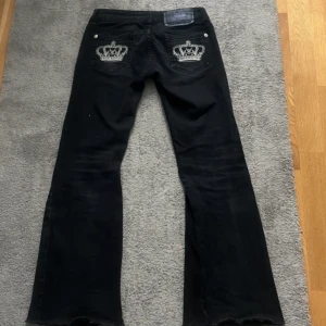 Viktoria Beckham jeans - Snygga svarta bootcut jeans med unika broderade kronor på bakfickorna. Jeansen har en klassisk femficksdesign och knappar i silverfärg. Perfekta för en stilren look med en twist.