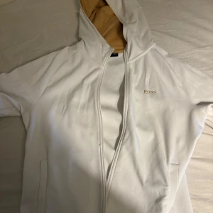 Vit hoodie från Boss - Säljer en stilren vit hoodie från Boss med dragkedja och logotyp på bröstet. Hoodien har en guldig insida i huvan och ribbade muddar. Perfekt för en avslappnad stil. Säljer denna otrolig sällsynta tjocktröja, som inte säljs längre! Priset kan diskuteras vid en snabbaffär och är även öppen för byten!