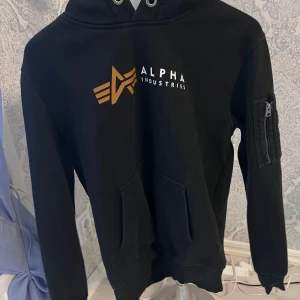 Svart hoodie från Alpha Industries - Säljer en svart hoodie från Alpha Industries med logotyp på bröstet och en praktisk ficka med dragkedja på ärmen. Perfekt för en casual look. 🖤