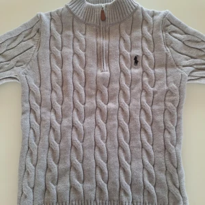 Grå stickad tröja från Polo Ralph Lauren - Säljer en stilren grå stickad tröja från Polo Ralph Lauren med kabelstickat mönster. Tröjan har en dragkedja vid halsen och en broderad logotyp på bröstet. Perfekt för kyliga dagar!