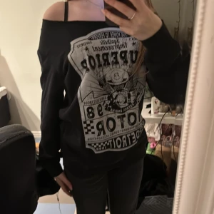 Svart offshoulder tröja med tryck - Säljer en svart offshoulder tröja med ett stort tryck på framsidan. Tröjan har en avslappnad passform och långa ärmar. Perfekt för en cool och avslappnad stil.