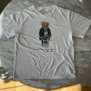 Vit t-shirt med Polo Bear från Ralph Lauren - Säljer en vit t-shirt från Ralph Lauren med Polo Bear-tryck på framsidan. Björnen är klädd i en stilren outfit med jeans och kavaj. Perfekt för en avslappnad stil. T-shirten är kortärmad och gjord i bomull. Den är XL men passar storleken M och har längden 63cm och bredden 50cm