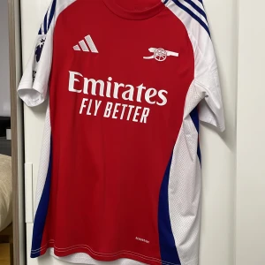 Röd fotbollströja från Adidas - Säljer en röd fotbollströja från Adidas med vita och blå detaljer. Tröjan har korta ärmar och tryck med texten 'Emirates Fly Better' på framsidan. På baksidan finns namnet 'Saka' och nummer 7. Premier League-logga och 'No room for racism' märke på ärmen. Storlek L men passar M
