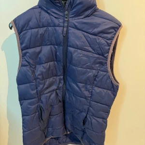 Blå dunväst från b.young - Säljer en blå dunväst från b.young med dragkedja framtill. Västen har en quiltad design och är perfekt för kyligare dagar. Den är ärmlös och har en hög krage för extra värme. Passar bra till lager-på-lager-stil.