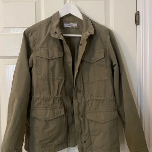 Grön jacka från Ellos - Säljer en grön jacka från Ellos i storlek 34. Jackan har en klassisk design med knappar och flera fickor framtill. Perfekt för vår och höst med sin stilrena look och praktiska detaljer.