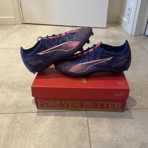 Puma Ultra 5 Pro FG/AG Fotbollsskor - Säljer ett par Puma Ultra 5 Pro FG/AG fotbollsskor i färgen Lapis Lazuli och vit. Skorna har en snygg design med snörning och är perfekta för både gräs och konstgräs. De har en blå och rosa färgkombination med en slitstark sula för optimalt grepp.