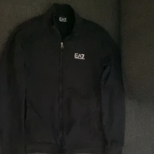 Svart tröja med dragkedja från Emporio Armani - Snygg svart tröja från Emporio Armani med dragkedja och EA7-logga på bröstet. Perfekt för en stilren look. Tröjan har långa ärmar och en hög krage för extra komfort.