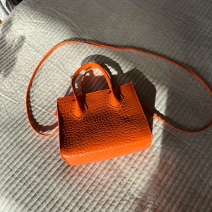 Orange axelväska i syntet - Snygg orange axelväska med krokodilpräglat mönster. Väskan har både handtag och en lång axelrem för flexibel användning. Perfekt för att ge en färgklick till din outfit! Ca 20x17 cm (LxH)