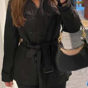 Svart trenchcoat från Orsay - Snygg svart trenchcoat från Orsay med knappar och bälte i midjan. Jackan har en klassisk krage och långa ärmar, perfekt för höst och vår. En stilren och tidlös design som passar till många tillfällen. Den är i storlek 42 men passar mig som vår S då den är liten i storleken 
