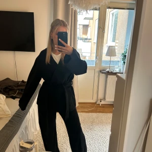 Svart kappa med bälte - Snygg svart kappa med bälte som ger en elegant look. Jackan har långa ärmar och en omlott-design som knyts i midjan. Perfekt för både höst och vår. Har även en knapp man kan knäppa utöver bältet. Helt oanvänd!!