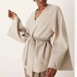 Beige kappa med bälte - Elegant beige omlottjacka med knytbälte i midjan. Jackan har vida ärmar och en stilren design som passar perfekt för både höst och vår. Den är tillverkad i ett mjukt material som ger en bekväm passform.