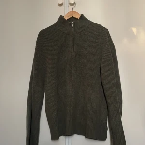 Olivgrön stickad tröja med dragkedja - Säljer en olivgrön stickad tröja med dragkedja i halsen. Tröjan har en ribbad struktur och långa ärmar, perfekt för kyligare dagar. Enkel och stilren design som passar till det mesta. 