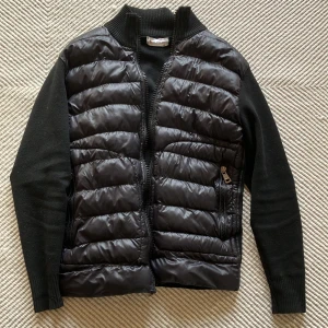 Svart cardigan/jacka från Moncler - Säljer en stilren svart cardigan jacka från Moncler. Jackan har långa ärmar och en dragkedja framtill. Den är perfekt för kyligare dagar och har en snygg ribbad krage och muddar. En klassisk och tidlös design för den modemedvetna.
