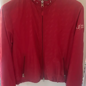 Röd vindjacka från Armani Jeans - Snygg röd vindjacka från Armani Jeans med dragkedja och huva. Jackan har långa ärmar och två fickor med dragkedja framtill. Perfekt för blåsiga dagar!