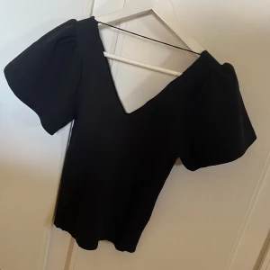Svart ribbad topp med volangärm  - Säljer en stilren svart ribbad topp med v-ringning och puffiga kortärmar. Perfekt för en trendig look. Passar till både byxor och kjol! Toppen är aldrig använd 