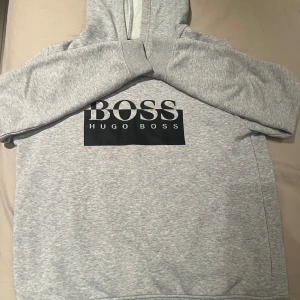 Grå hoodie från Hugo Boss - Säljer en stilren grå hoodie från Hugo Boss med deras ikoniska logga tryckt på framsidan. Tröjan har långa ärmar och en klassisk huva. Perfekt för en avslappnad look. Hör av er om ni behöver fler bilder🙌