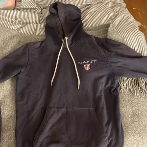 Mörkblå hoodie från GANT - Säljer en snygg mörkblå hoodie från GANT med broderad logga och emblem på bröstet. Tröjan har en justerbar huva med vita snören och en praktisk magficka. Perfekt för en avslappnad stil.