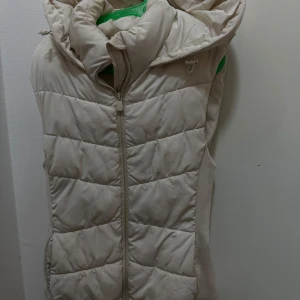 Beige quiltad dunväst med huva - Säljer en snygg beige quiltad dunväst med dragkedja och huva från Zara. Den har en sportig look och är lätt att matcha med olika outfits.