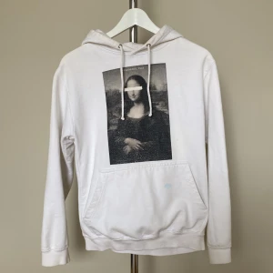 Vit hoodie med Mona Lisa-tryck - Vit hoodie med ikoniskt Mona Lisa-tryck på bröstet och klassisk känguruficka. Tröjan har huva med snörning och långa ärmar. Perfekt för dig som gillar streetstyle och konstnärliga detaljer. Har en blå fläck (se bild 2). 