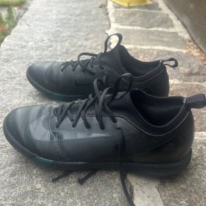 Svarta Nike fotbollsskor med snörning - Snygga svarta Nike fotbollsskor med diskret Swoosh-logga på sidan. Skorna har snörning, låg profil och är tillverkade i syntetmaterial med texturerad yta för bättre bollkontroll. Sulan är mönstrad och har detaljer i mörkgrönt. Perfekta för dig som vill ha stilrena skor på planen.