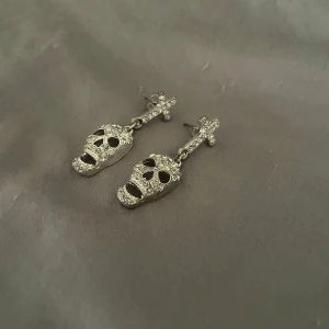Glittriga döskalleörhängen i silver - Säljer ett par coola örhängen i silverfärg med glittrande stenar. Hängande modell med döskalleform och kors upptill.