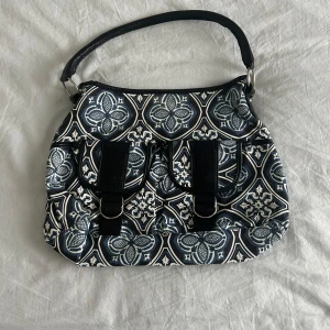 Mönstrad handväska från Complement - Snygg handväska med blått och vitt grafiskt mönster, två fickor framtill med spännen och magnetlås. Väskan har svart axelrem och innerfack med dragkedja. Perfekt för dig som vill sticka ut med en unik accessoar. Aldrig använd, nyskick!