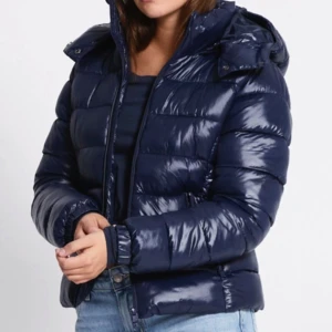 Mörkblå pufferjacka med huva - Snygg mörkblå pufferjacka med glansig finish och huva. Jackan har quiltad design, dragkedja framtill och är perfekt för kalla dagar. Passformen är normal och jackan är lätt att matcha med jeans eller andra byxor.