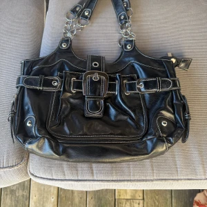 Svart handväska från Snooke med kedjedetaljer - Cool svart handväska från Snooke i skinnimitation med silvriga kedjor på axelbanden och snygga spänndetaljer framtill. Väskan har vita kontrastsömmar, dragkedja upptill och flera fack för småprylar. Perfekt för dig som gillar statement-accessoarer.