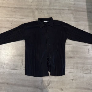 Svart plisserad skjorta från Kappahl - Svart skjorta från Kappahl i storlek S med plisserat tyg och klassisk krage. Skjortan har lång ärm och knäppning framtill. Materialet är lätt och ger en snygg struktur. Perfekt för dig som gillar stilrena och moderna plagg.