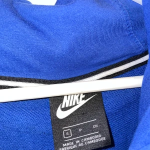 Blå Nike hoodie med dragkedja - Blå hoodie från Nike med hel dragkedja, huva med snörning och kängurufickor. Liten röd Nike-logga broderad på bröstet. Skön och sportig stil, perfekt för chill eller träning.