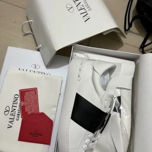 Valentino Garavani Open sneakers vit/svart - Snygga Valentino Garavani Open sneakers i vitt skinn med en bred svart rem över mitten. Klassisk rund tå, platt sula och diskreta nitar på hälen. Perfekta för dig som vill ha en stilren och lyxig look med en modern twist.