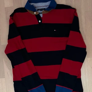 Randig pikétröja Tommy Hilfiger - Snygg pikétröja från Tommy Hilfiger med breda röda och marinblå ränder, klassisk krage i blått och bruna detaljer vid knäppningen. Liten logga på bröstet och långa ärmar. Perfekt för en sportig och avslappnad look.