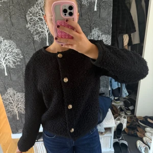 Svart kofta med guldknappar - Mysig svart kofta med fluffig, stickad känsla och snygga guldknappar framtill. Rund halsringning och långa ärmar. Perfekt att slänga på sig när det är kyligt ute och vill ha något både stilrent och bekvämt.