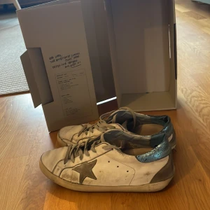 Golden Goose vita sneakers med blå detalj - Säljer ett par vita golden goose med blå detaljer i dåligt skick(därav priset) en defekt på högra skon. Box medföljer vid köp!