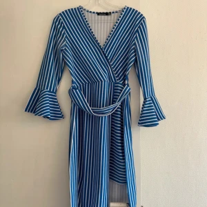 Blåvit randig omlottklänning Boohoo - Snygg blå och vit randig omlottklänning från Boohoo med v-ringning och volangärmar. Klänningen har knyt i midjan och trekvartsärm, vilket ger en cool och trendig look. Perfekt för dig som gillar färg och mönster!