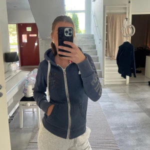 Mörkblå hoodie från Superdry - Mysig mörkblå hoodie från Superdry med dragkedja framtill, vita snören i huvan och tryck på bröstet och ena ärmen. Två fickor framtill och ribbade muddar. Perfekt för chill dagar eller när du vill vara extra bekväm.