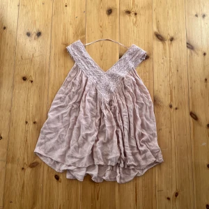 Ljusrosa blus med spetsdetalj - Supersöt ljusrosa blus med v-ringad spets upptill och luftigt, vågigt tyg. Perfekt för varma dagar när du vill ha något lätt och flowy. Blusen är ärmlös och har en romantisk vibe med sin breda spetskant och avslappnade passform.