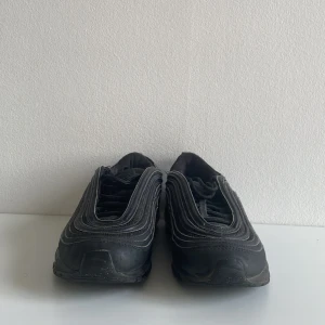  Nike Air Max 97  - Svarta Nike Air Max 97 med ikoniska vågformade linjer och synlig Air-sula. Skorna har snörning och en sportig vibe, tillverkade i syntet och textil. Perfekta för dig som gillar streetstyle och vill ha en klassisk sneaker med modern känsla.