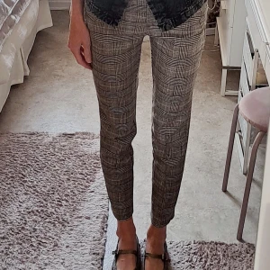 Rutig croppad kostymbyxa - Snygga kostymbyxor från HM med rutig mönster i bruna toner. Byxorna är croppade och har en slim passform som sitter tajt längs benen. De har klassisk midja och är perfekta för dig som gillar stilrena och trendiga plagg. Storlek 36, men mer som en 34. Liten i midjan och över höfter. 