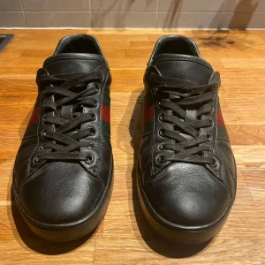 Gucci svarta sneakers med röd/grön rand - Snygga svarta sneakers från Gucci i skinn med klassiska rödgröna ränder på sidorna och röd detalj vid hälen. Skorna har svarta snören, rund tå och platt sula med Gucci-logga undertill. Perfekta för dig som vill ha en stilren och lyxig look.